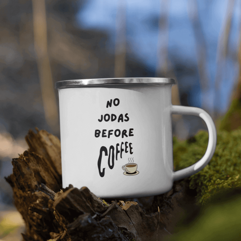 A durable Ecomartists enamel mug with the phrase "no jodas before coffee" printed on it, resting on a wooden stump outdoors, perfect for your outdoor adventures.
