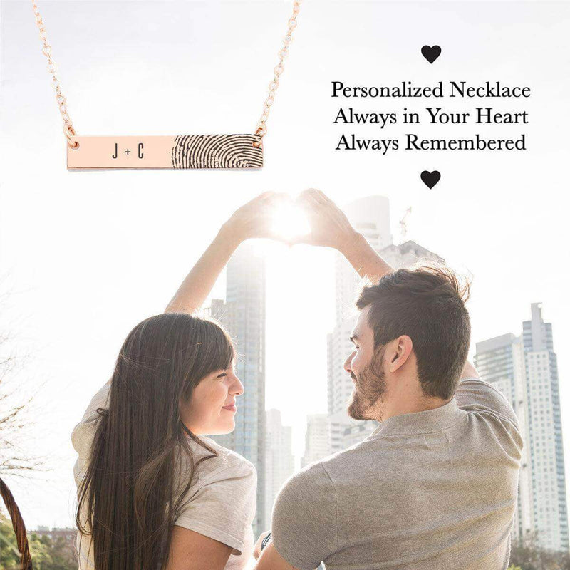 Couple forming a heart shape with their hands, showcasing an Ecomartists Fingerprint Bar Necklace with Custom Engraving and skyscrapers in the background.